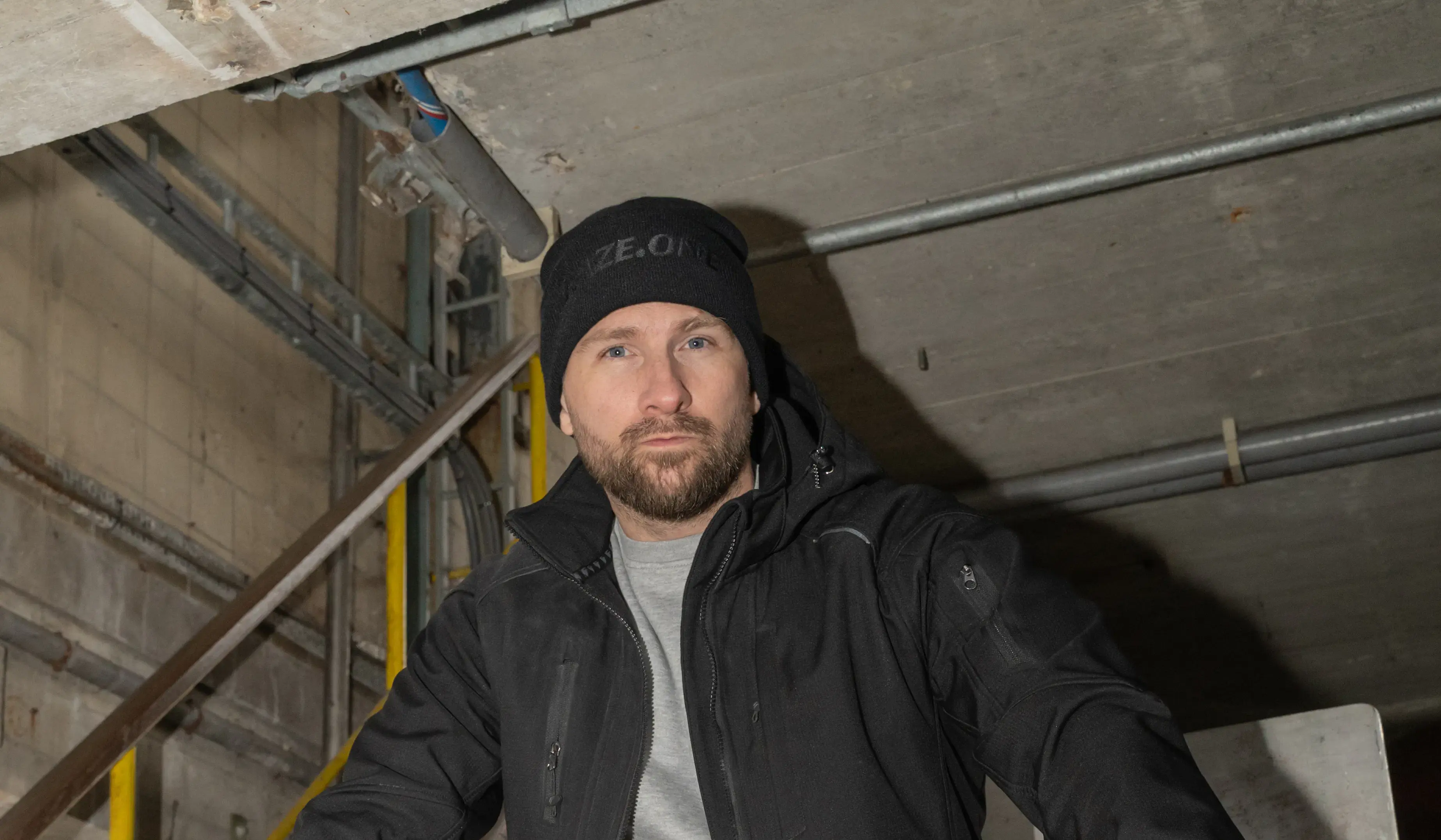 Man wearing black Faze.One jacket and beanie on an industrial staircase, casual workwear style